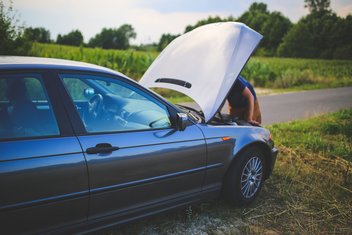 reparation-car-accident.jpg A man is repairing a car, with the hood open, on the side of a country road.