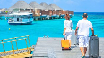 travel-package-beach-hotel.jpg A couple walking suitcases by hand on a footbridge over turquoise water. Wooden and straw bungalows can be seen in the background.