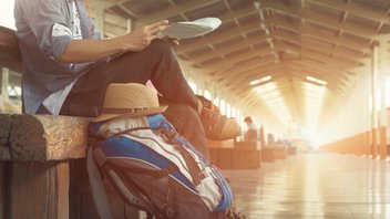 A person sitting on a bench waits at a train station, their travel bag placed beside them.