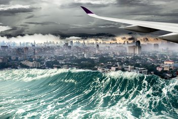 The wing of an aircraft flies over a tsunami wave crashing into a city